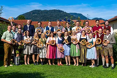Group photo of some employees in Bavarian costume and wearing a large pretzel in the garden of the itelio GmbH headquarters in Kiefersfelden with a view of the Nusslberg.
