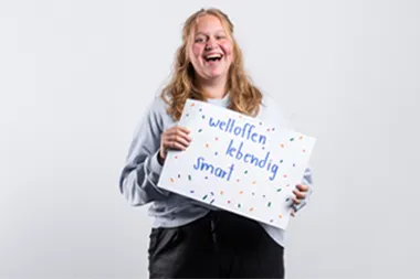 Eine Auszubildende hält ein selbst gestaltetes Schild mit den Worten weltoffen, lebendig, smart in den Händen, um die Unternehmenskultur von itelio zu beschreiben.