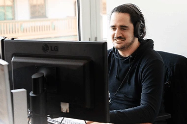 Ein junger Mitarbeiter der itelio GmbH sitzt lächelnd vor einem Monitor.